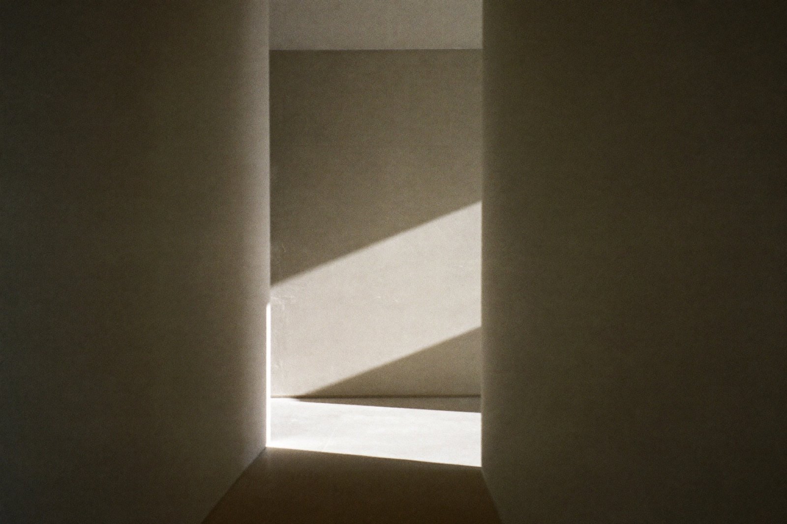 A bare pale plaster corridor with a diagonal shaft of low sunlight crossing the wall and floor.