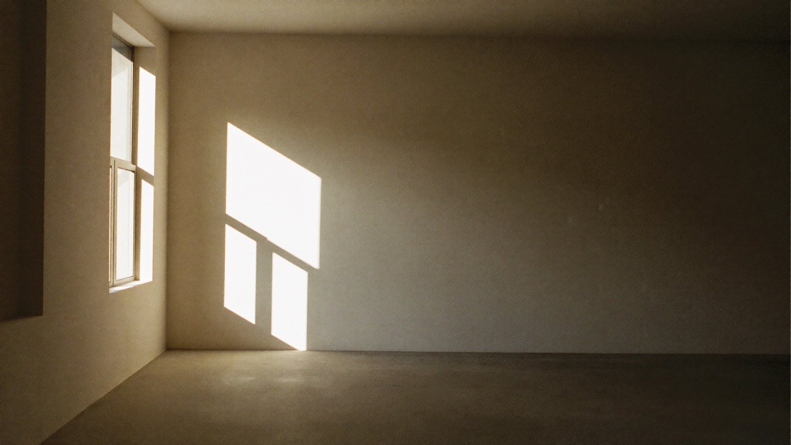 An empty room with bone-white plaster walls, lit by a single hard rectangle of afternoon sunlight from a window.
