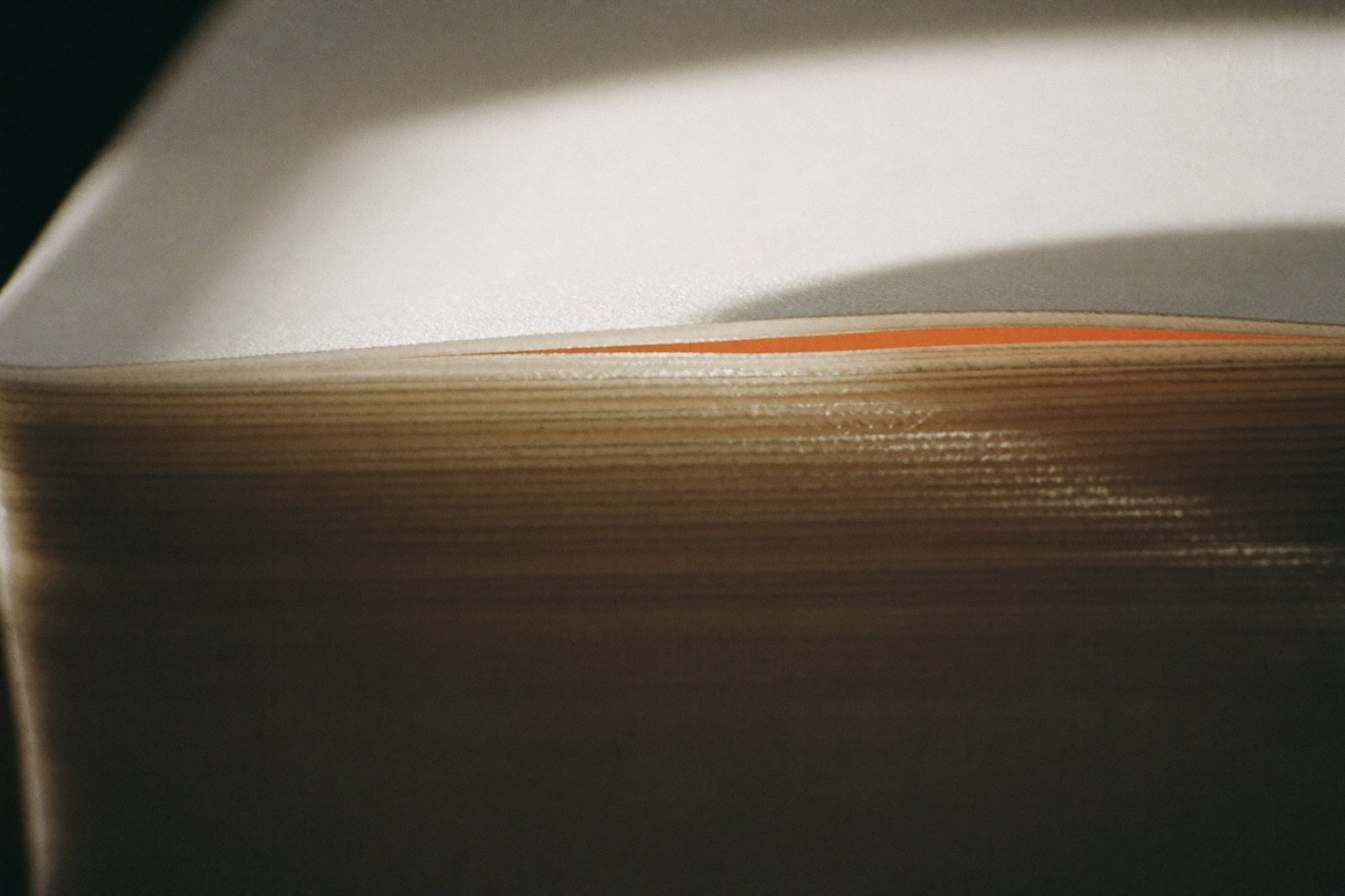 Close-up of the cut edge of a stack of thick matte paper, raking sidelight picking out the layers, one sheet tinted burnt orange.